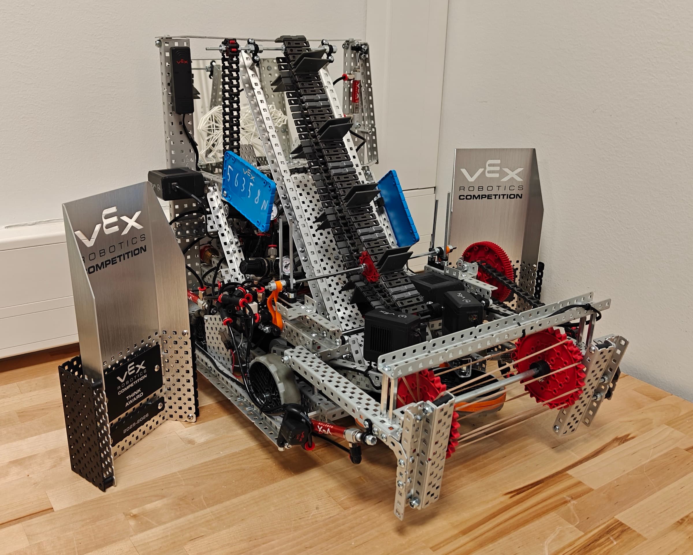 Our VEX V5 competition robot on the bench: intake rollers at the front, the chain conveyor climbing to the tower behind them, license plates on both sides.